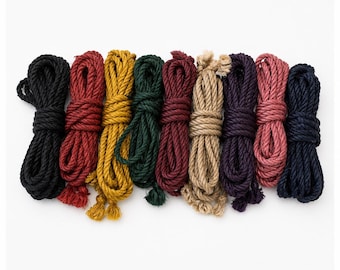 Op de afbeelding: Een verzameling van negen opgerolde touwen in verschillende kleuren: zwart, rood, geel, groen, bordeaux, bruin, paars, roze en marineblauw. De touwen zijn horizontaal gerangschikt tegen een witte achtergrond, wat hun textuur en kleurvariaties benadrukt.