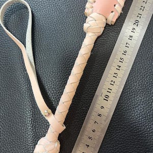 May include: A pink and white leather whip with a braided handle and a loop for holding. The whip is measured against a ruler, showing it is approximately 25 inches long.