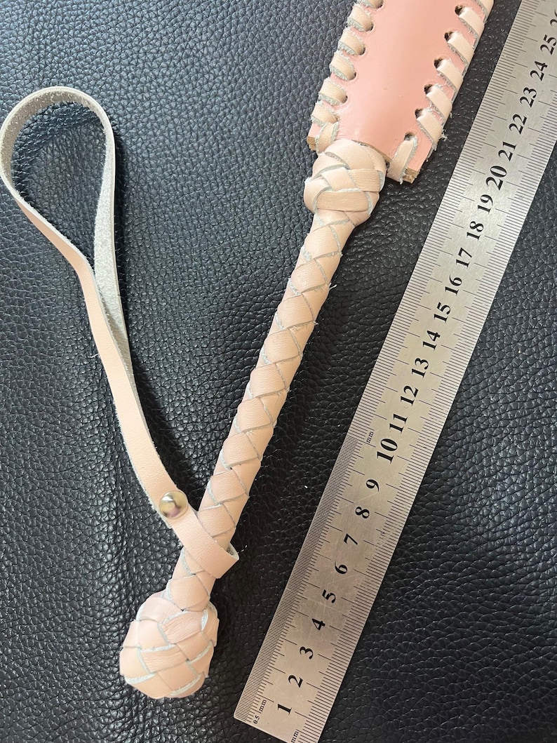 May include: A pink and white leather whip with a braided handle and a loop for holding. The whip is measured against a ruler, showing it is approximately 25 inches long.