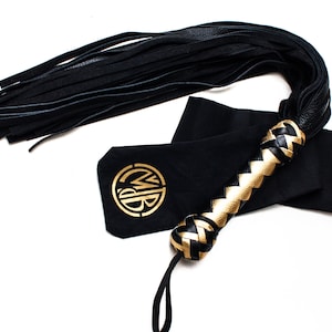 May include: Black leather flogger with a braided handle featuring gold accents. The flogger is accompanied by a black pouch with a gold logo. The design includes a long fringe of black leather strips.