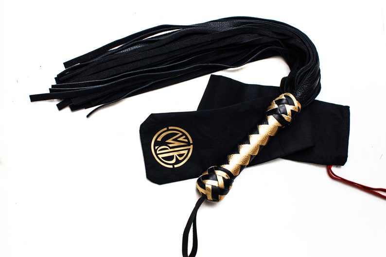 May include: Black leather flogger with a braided handle featuring gold accents. The flogger is accompanied by a black pouch with a gold logo. The design includes a long fringe of black leather strips.