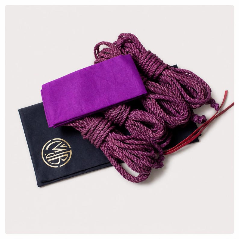 May include: A set of purple braided ropes coiled neatly alongside a folded purple fabric rectangle and a black rectangular pouch featuring a circular gold logo with the letters MB in the center, accompanied by two thin red leather strips.