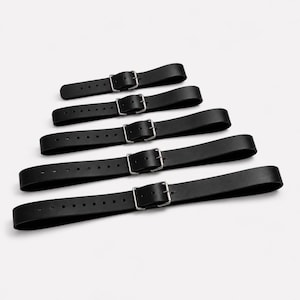 Peut inclure: Cinq lanières en cuir noir disposées par ordre de longueur croissante. Chaque lanière présente une série de trous perforés pour le réglage et une boucle en métal argenté. Les lanières ont une texture lisse et mate, disposées horizontalement.
