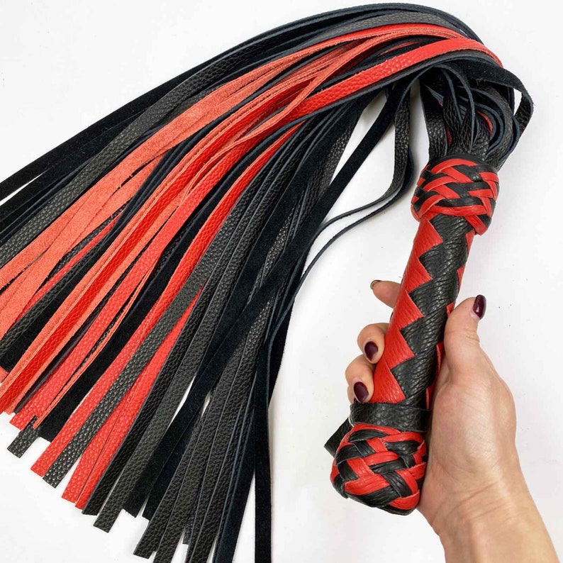 May include: A black, red, and black leather whip. The handle is woven in a red and black pattern. The whip's strands are a mix of black and red leather strips. The whip is held in a hand.