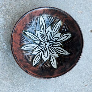 May include: A handmade ceramic bowl with a floral design. The bowl features a dark brown rim and a lighter brown interior. A white floral pattern is centered in the bowl. The bowl is round and sits on a gray surface.
