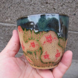 May include: A ceramic cup with a speckled tan base featuring red bee illustrations and green drip glaze accents. The upper rim is finished with a dark blue-green glaze that flows down over the tan surface, creating a textured, multi-colored appearance.