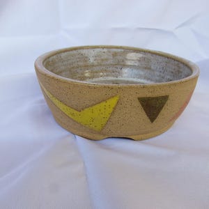 May include: A speckled, tan-colored ceramic bowl with a wide rim. The bowl features yellow, brown, and pink geometric shapes. The interior is a lighter tan color. The bowl is likely handmade and suitable for serving food or as a decorative piece.