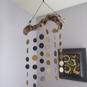 May include: A hanging mobile featuring a natural driftwood branch suspended from the ceiling. Strands of circular ceramic discs in shades of blue, tan, and cream dangle below. A framed abstract painting hangs on the wall.