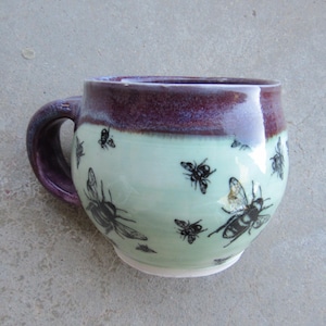 May include: A ceramic mug with a rounded body featuring a light mint green base color and a dark purple glazed rim. The exterior is decorated with multiple black bee illustrations of varying sizes scattered across the mint green surface.