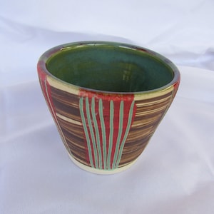 May include: A handmade ceramic bowl with a unique design. The bowl features a brown exterior with red and green vertical stripes. The interior is a deep green color, and the rim has a red accent. The bowl is set against a white background.