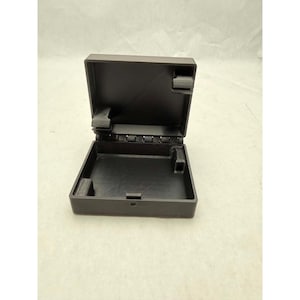May include: A matte black square storage box with a hinged lid. The interior features a flat base with small molded compartments in the corners and a central hinge mechanism connecting the lid to the base, providing a secure enclosure.