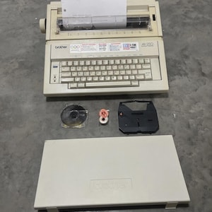 May include: Off-white Brother AX250 typewriter with a full keyboard and paper inserted. Includes a cover, ribbon, and other accessories. The typewriter has the Brother logo and Olympic rings on the front.