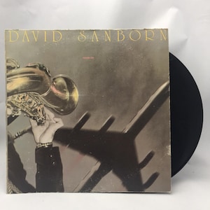 Könnte beinhalten: Vintage Vinyl-Schallplattenhülle für David Sanborns "Taking Off". Das Cover zeigt ein Saxophon, eine Hand und einen Schatten. Der Albumtitel und der Name des Künstlers sind in goldfarbenen Buchstaben gehalten. Die Schallplatte selbst ist schwarz.