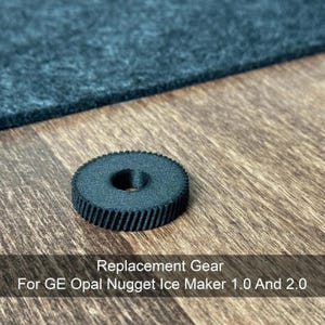 Op de afbeelding: Een zwarte vervangende tandwiel voor GE Opal Nugget Ice Maker 1.0 en 2.0. Het tandwiel is cirkelvormig met een centraal gat en geribbelde randen. Het tandwiel ligt op een houten oppervlak met een donkerblauw viltachtig materiaal op de achtergrond. Tekst op de afbeelding luidt "Replacement Gear For GE Opal Nugget Ice Maker 1.0 And 2.0."
