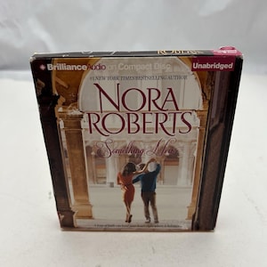 May include: A compact disc of Nora Roberts' "Something New" audiobook. The cover features a couple in front of a building. The title and author's name are prominently displayed. The packaging includes "Brilliance Audio" and "Unabridged" text.