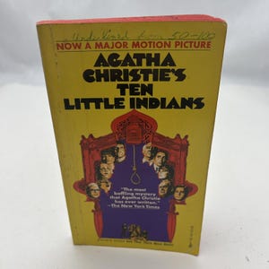 以下が含まれることがあります： 「アガサ・クリスティの10人のインディアン」というタイトルのヴィンテージペーパーバック。表紙は黄色で、黒い文字と紫と赤の顔のイラストが描かれています。本の上の端は赤で、「Now a Major Motion Picture」と書かれています。