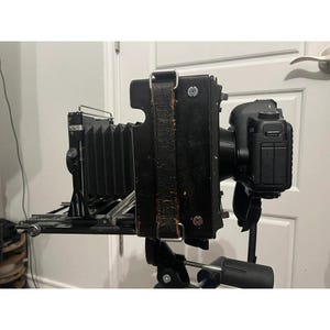 May include: A vintage black large format camera with a bellows design, mounted on a tripod. The camera has a leather strap and is paired with a modern digital camera body. The setup is designed for professional photography.