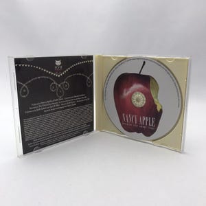 Puede incluir: Una caja de CD de plástico abierta que presenta un inserto negro con texto blanco y un logotipo de gato a la izquierda. El lado derecho contiene un disco con el gráfico de una manzana roja mordida, etiquetado como Nancy Apple Shoulda Lied About That.