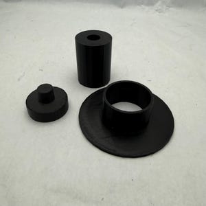 May include: Three black 3D-printed objects. A cylindrical piece with a hole in the center, a small disc with a raised cylinder, and a flat disc with a raised ring. The objects are made of a matte black material.