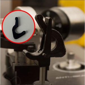 Puede incluir: Primer plano de un componente negro en forma de gancho, impreso en 3D, posiblemente parte de un mecanismo más grande. El gancho está dentro de un círculo rojo, con una estructura negra más grande en el fondo. La imagen se centra en los detalles del gancho.