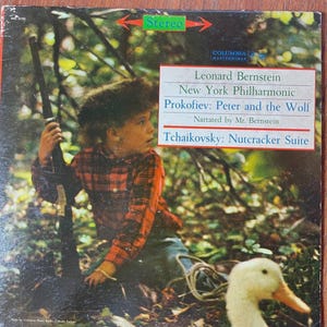 Può includere: Copertina di un album in vinile vintage con Leonard Bernstein e la New York Philharmonic. L'album include "Pierino e il lupo" di Prokofiev e "Lo schiaccianoci" di Tchaikovsky. La copertina mostra un bambino con un fucile e un'anatra.