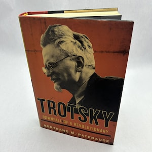 May include: A hardcover book titled "Trotsky: Downfall of a Revolutionary" by Bertrand M. Patenaude. The cover features a sepia-toned portrait of a man wearing glasses, with the title in bold, orange letters against an orange background.