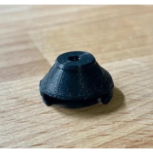May include: A black, 3D-printed object with a conical shape and a circular top. The object has a textured surface and appears to be a small, functional component, possibly a cap or a stopper. The object is sitting on a wooden surface.