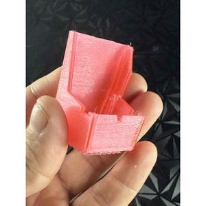 May include: A small, coral-colored 3D-printed object with a rectangular base and angled sides. The object has a series of parallel lines and ridges, suggesting a layered construction. The item is held in a hand against a dark, textured background.