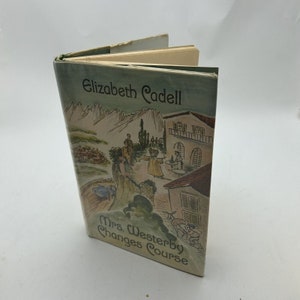 May include: A vintage book titled "Mrs. Westerby Changes Course" by Elizabeth Cadell. The cover features a watercolor illustration of a European village scene with mountains, buildings, and figures. The book is open, revealing aged, cream-colored pages.
