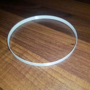 Puede incluir: Un objeto blanco de forma circular, posiblemente un anillo o una banda, está centrado en la imagen. El objeto está hecho de un material sólido y opaco. El fondo es una superficie de madera de color marrón.