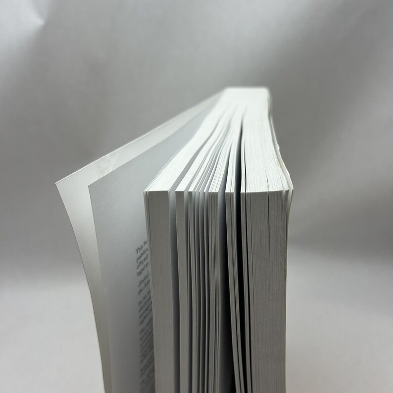 K&ouml;nnte beinhalten: Ein Hardcover-Buch mit wei&szlig;en Seiten, von der Seite betrachtet. Die Seiten sind leicht aufgef&auml;chert und geben den Text preis. Der Buchr&uuml;cken ist nicht sichtbar, und der Hintergrund ist ein schlichtes, hellgraues Grau.