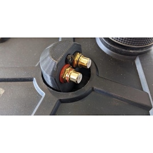 May include: A black 3D-printed component featuring two gold-plated RCA connectors with red and black rubber gaskets, mounted into a circular opening on a dark gray textured surface, secured by a small black hex screw on the top side.