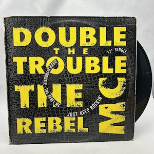May include: A black and yellow vinyl record cover with the text "Double The Trouble The Rebel MC Just Keep Rockin'" on it. The record is a 12" single.