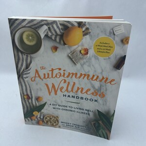 May include: The Autoimmune Wellness Handbook, a DIY guide to living well with chronic illness. The cover features orange lettering and includes a 4-Week Meal Plan and a 12-Week Lifestyle Plan. A cup of tea and a lemon are visible.