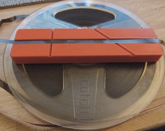 May include: A rectangular orange plastic tape splicing block with diagonal and straight grooves, positioned horizontally across a circular metal Scotch brand reel containing dark magnetic recording tape.