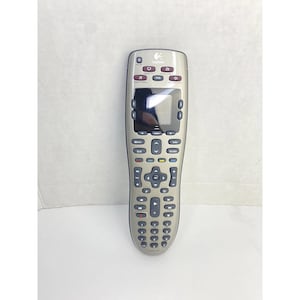 Op de afbeelding: Een zilveren Logitech afstandsbediening met een zwart scherm en talrijke knoppen. De afstandsbediening heeft een gebogen ontwerp en beschikt over knoppen voor aan/uit, volume en kanaalbediening. Het Logitech-logo is zichtbaar bovenaan.