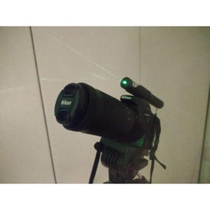 May include: A black Nikon camera with a lens cap and a green laser pointer attached. The camera is mounted on a tripod, and a green laser beam is visible, extending from the pointer.