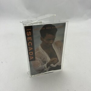May include: A clear cassette tape case with a cover featuring a man in a wet white shirt. The text "Jon Secada" is visible on the side, with the album title "Otro dia mas sin verte" on the front.