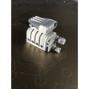 May include: A gray 3D-printed model of an engine, featuring a rectangular air intake and multiple curved cylinders. The model has detailed textures and a mechanical design, showcasing the intricate components of an engine.