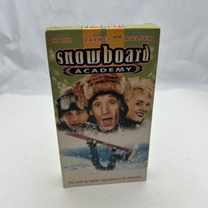 Puede incluir: Una cinta VHS vintage de la película "Snowboard Academy". La portada presenta a Corey Haim, Jim Varney y Brigitte Nielsen. El título está en negrita, con una combinación de colores verde y rojo. El lema dice: "El camino hacia la educación superior es todo cuesta abajo."