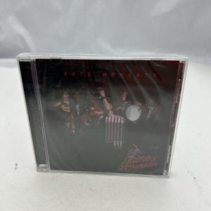 Peut inclure: Un boîtier de CD scellé avec une image de groupe sur fond sombre. Le texte "ESTE MOMENTO" est en haut, avec "Jaime y Chamacos" en cursive en bas. Les membres du groupe sont visibles à travers l'emballage plastique.