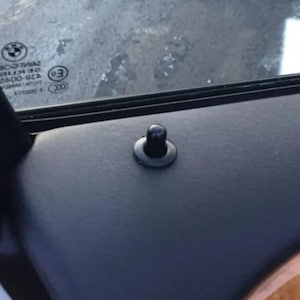 Puede incluir: Primer plano de un pasador de bloqueo de puerta de coche negro. El pasador es cilíndrico con una parte superior redondeada y una base circular. La superficie circundante es gris oscuro.