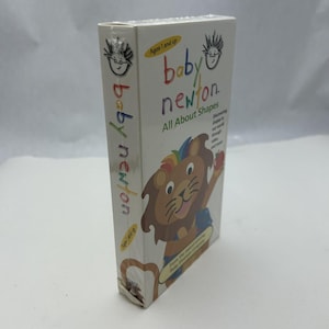 May include: A white Baby Newton VHS tape box. The box features a cartoon lion with a rainbow mane and the text "Baby Newton All About Shapes". The box is for ages 1 and up.