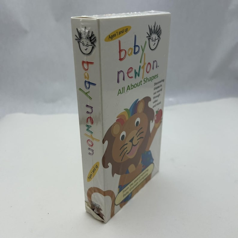 May include: A white Baby Newton VHS tape box. The box features a cartoon lion with a rainbow mane and the text "Baby Newton All About Shapes". The box is for ages 1 and up.