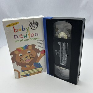 May include: A Baby Newton VHS tape and its packaging. The packaging features a lion illustration with a rainbow mane and the text "All About Shapes". The VHS tape is black with a blue spine.