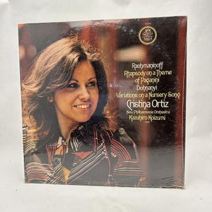 May include: A vintage vinyl record album cover featuring a woman with brown hair and a red and black striped shirt. The album is titled "Rachmaninoff Rhapsody on a Theme of Paganini Dohnanyi Variations on a Nursery Song" and features the performer "Cristina Ortiz" with the "New Philharmonia Orchestra" conducted by "Kazuhiro Koizumi".