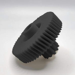 May include: A black gear with interlocking teeth, featuring a central hole. The gear is angled, showcasing its detailed design and mechanical structure. The object is set against a plain white background.