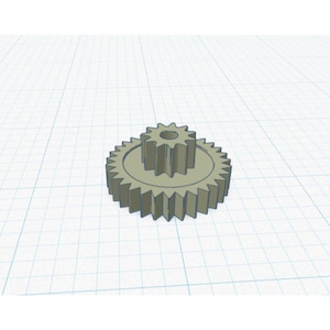 May include: A light beige gear assembly with two interlocking gears. The larger gear has a circular base with evenly spaced teeth around the perimeter. The smaller gear sits atop the larger one, also with teeth. The background is a grid.