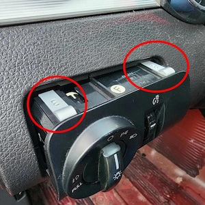 May include: Close-up of a car's dashboard components. The image shows a black control panel with a rotary switch and several rectangular, gray buttons. The buttons are labeled with white text. The dashboard has a textured, dark gray surface.