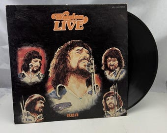 Könnte beinhalten: Ein schwarzes Vinyl-Schallplattencover mit einem Foto von Waylon Jennings in der Mitte. Der Albumtitel lautet "Waylon Live" und das Plattenlabel ist RCA. Das Albumcover zeigt mehrere Fotos von Waylon Jennings in verschiedenen Posen.